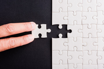 Top view flat lay of hand-holding last piece white paper jigsaw puzzle game last pieces put to place for solve problem complete mission, studio shot on a black background, quiz calculation concept