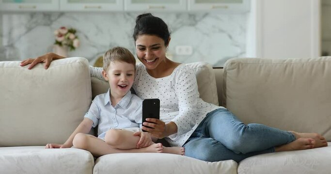 Indian babysitter and Caucasian little boy having fun using new cool mobile application, sit together on sofa at home looking at smartphone screen enjoy on-line e amusements, modern tech usage concept