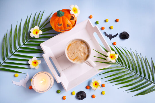 Cup of coffee, orange pumpkin, burning candle, bats, candies and tropical palm leaves on the blue background. Halloween on the exotic island. Coffee time on the beach. Tropical Halloween concept.