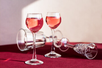 a pair of glasses of red alcoholic drink among empty glasses lying on the table, light and shadow on the background, rose berry liqueur or cocktail among fallen tableware, elegant minimalism