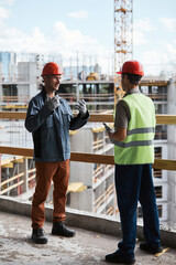 Vertical full length portrait of two construction workers discussing project while standing at high rise building