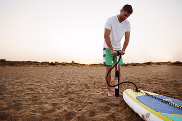 Young man inflating paddle sup board on the beach at sunrise