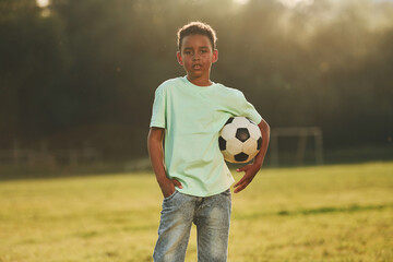 Sits with soccer ball. African american kid have fun in the field at summer daytime