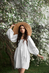 Brown-haired woman in a large straw hat and a white dress poses against the background of blooming white trees. Romantic look, natural beauty, clean facial skin