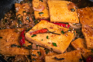 Fried hot tofu - chunks of fried tofu cheese in a hot sauce with sesame seeds, onions and peppers, close-up.