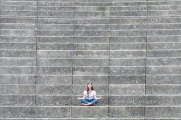 Casual dressed young woman sitting on stairs of a grandstand and meditating. Mental health, mindfulness and yoga concept with copy space