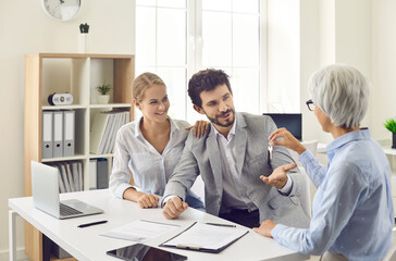 Obraz premium Senior realtor gives key to new house or apartment to happy young married couple after they have signed agreement. Concept of real estate, mortgage, rent, lease, or buying your own property