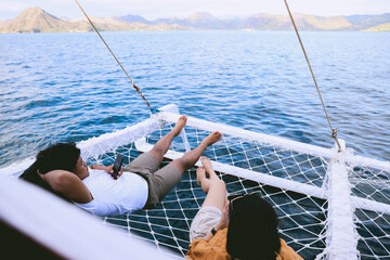 Asian couple relaxing on catamaran net while using smart phone with seascape and hills at Labuan Bajo