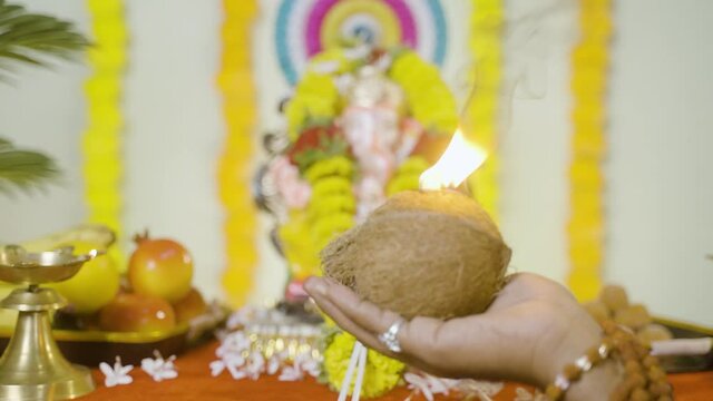 Rack focus from hands to idol, close up hands offering coconut aarti to lord ganesha during vinayak festival celebration at home.