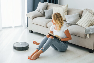 Blonde sits on the floor. Woman in casual clothes is indoors at home
