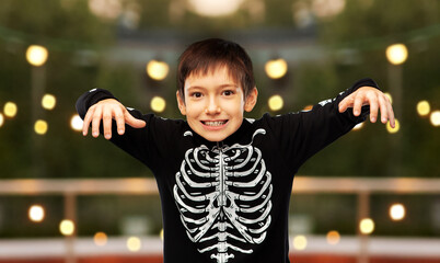 halloween, holiday and childhood concept - boy in black costume of skeleton frightening over...