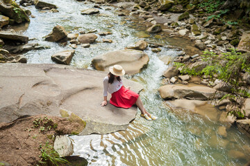 Stylish girl in a straw hat, white blouse, red skirt and yellow sneakers sitting above mountain river. Young woman enjoys fresh air in forest resting on a cliff near a waterfall. lonely travel.