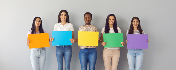 Banner background with group portrait of happy smiling young multiracial women standing together by white grey studio wall holding colorful orange, blue, yellow, green and purple mockup paper signs