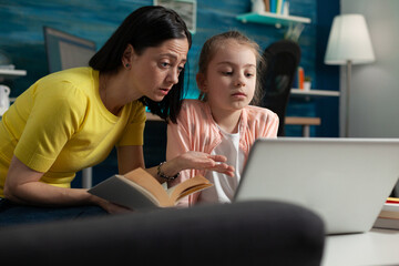 Mother and daughter doing homework for online school class with teacher sitting at home desk. Adult parent giving education knowledge to little child preparing for lesson lecture on laptop