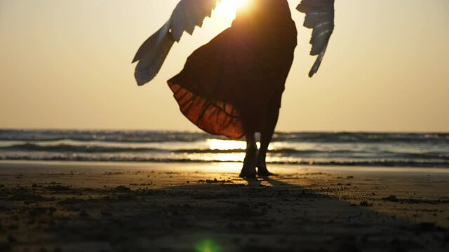 Silhouette of beautiful female angel walking barefoot from camera toward the sea at sunset, rear view