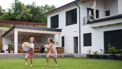 Two small girls sisters playing with a ball outdoors in the backyard, running. - obrazy, fototapety, plakaty