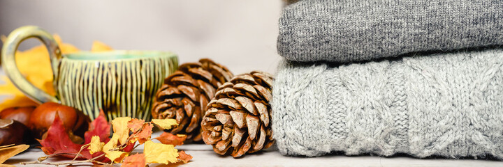 autumn yellow maple leaves, pine cones, chestnuts and a cup of tea next to a stack of knitted cozy warm winter sweaters in several shades of gray on a gray background. fall concept. banner