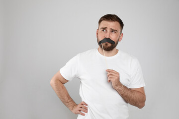Thoughtful man with fake mustache on light grey background, space for text