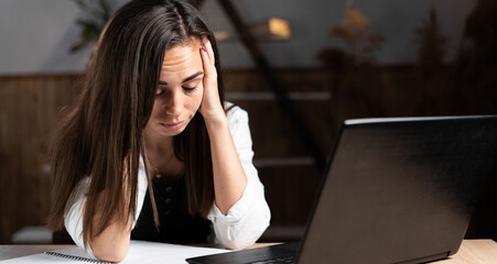 Sad tired Caucasian woman sitting with laptop in office or at home under stress from bad news. girl holds her head with her hands unmotivated employee at the workplace. Work and health concept.