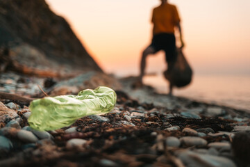 Close up of dirty green plastic bottle lies on the beach. Defocused silhouette of a person holding...