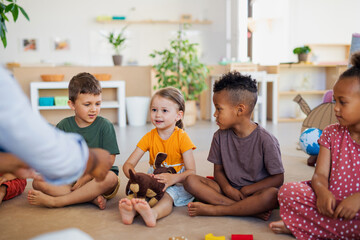 Group of small nursery school children sitting on floor indoors in classroom, listening to teacher.