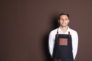 Portrait of happy young waiter in uniform on brown background, space for text