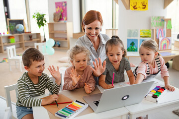 Group of small nursery school children with teacher indoors in classroom, using laptop. - obrazy, fototapety, plakaty