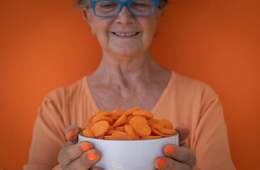 A mature woman holds up a bowl full of sliced carrots ready to eat. Orange background