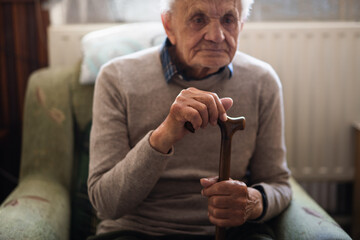 Sad elderly man with walking stick sitting on armchair indoors at home, resting.