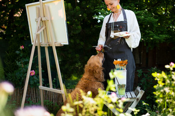 Young female artist working on her art canvas painting outdoors in her garden with golden retriever...
