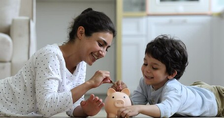Young Indian mother her little 4s son lying on floor at home count and drop coins into piggy bank. Caring parent teaches child save money, think about future, manage personal finances, savings concept