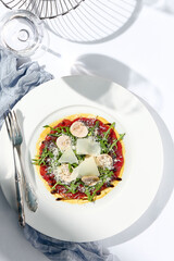 Beef carpaccio with mushrooms and parmesan cheese. Fancy dinning with beef carpaccio on white table with simple contemporary decor. Sunlight and harsh shadow still life. Beef carpaccio appetizer dish.
