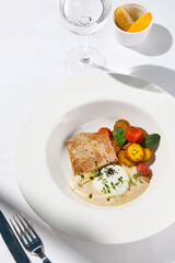 Fish steak with fillet tuna, warm tomato, baked potato and vitello tonnato sauce. Fish in white plate. Tuna sauce and vegetables. Contemporary seafood restaurant menu, summer day with sunlight shadow.