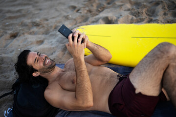 Handsome man resting on the beach, using the phone. Surfer taking a break on the beach..
