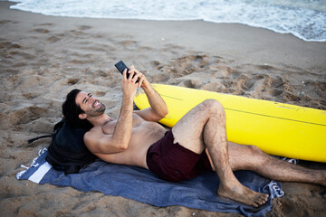 Handsome man resting on the beach, using the phone. Surfer taking a break on the beach..