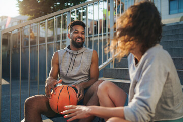 Man and woman friends with basketball resting after exercise outdoors in city, talking.