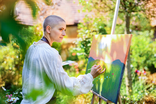 Beautiful young woman relaxing while painting an art canvas outdoors in her garden. Cancer survivor, mindfulness, art therapy, creativity concept.