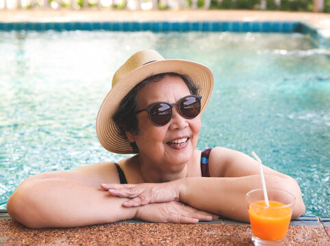 happy and healthy Asian senior  woman wearing straw hat and sunglasses  drinking  orange juice  in the swimming pool, smiling and looking away.