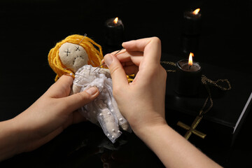 Woman stabbing voodoo doll with pin at black table, closeup. Curse ceremony
