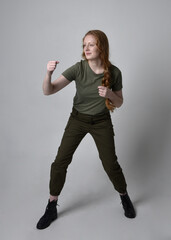 Full length portrait of pretty red haired woman wearing army green khaki shirt, utilitarian pants and boots. Standing pose isolated on studio background.