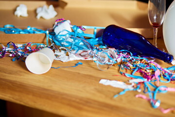 Messy wooden table with pizza box, empty blue bottles, wine glass and party decorations at morning...