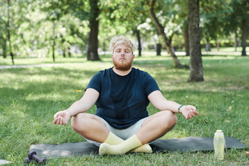 Naklejka na ściany i meble Overweight man sitting on the grass with eyes closed and meditating after sports training in the park