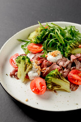 Healthy salad made from meat, cherry tomatoes, broccoli, goat cheese and fresh arugula served in a white plate.