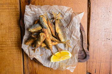 Small fried fish with a piece of lemon served on a rustic wooden board over wooden background.