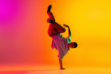 Portrait of young stylish man, break dancing dancer training in casual clothes isolated over gradient pink yellow background at dance hall in neon light.