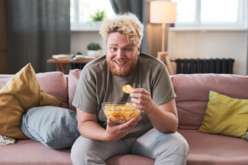 Overweight man sitting on sofa with bowl of chips eating them and laughing while watching TV at home