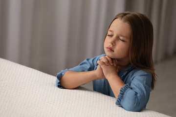 Cute little girl with hands clasped together saying bedtime prayer in bedroom. Space for text