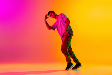 Full length portrait of young stylish man, break dancing dancer in casual clothes isolated over gradient pink yellow background at dance hall in neon light.