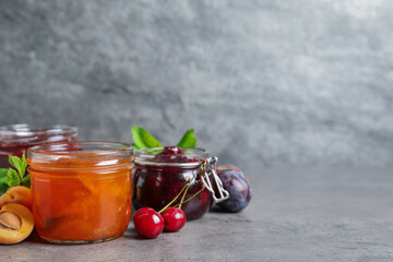 Jars with different jams and fresh fruits on grey table. Space for text