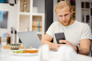 Bearded blonde man using cellphone and laptop while sitting on table